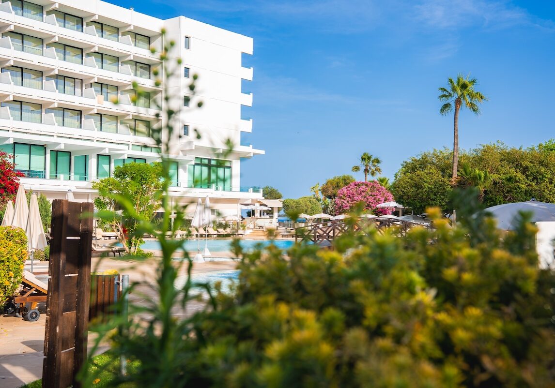 Modern white multi story hotel with balconies, a pool area, lush greenery, flowering bushes, palm trees, and a clear blue sky in Cyprus.